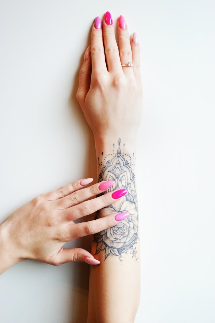 A close-up of tattooed arm and manicured hands with vibrant nail polish on a light background.